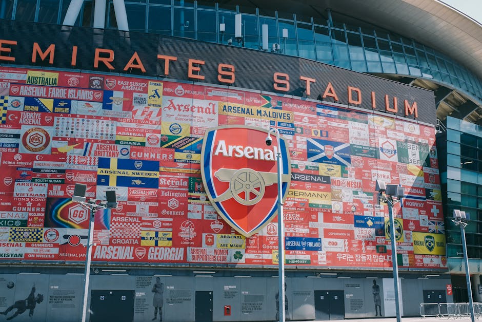 A large exterior wall of the Emirates Stadium, home to Arsenal Football Club, features a colorful mosaic mural composed of numerous individual tiles, each displaying various flags, symbols, and text related to Arsenal supporters from different countries and regions. The mosaic showcases vibrant reds, blues, yellows, and whites, with some tiles depicting the club’s logo and supporter slogans. Central to the mural is a prominent Arsenal crest, with a depiction of a cannon, set against a background of support banners. In front of the mural, three tall, metal-scaffolded floodlights with black rectangular fixtures stand on a paved ground, evenly spaced to illuminate the display area and surrounding space. The stadium’s modern architecture includes a curved, glass-paneled upper section and a sleek black facade with the stadium name partially visible at the top. Natural daylight illuminates the scene uniformly, highlighting the detailed textures of the tiles and the reflective quality of the glass facade, creating a professional and factual representation aligned with professional waste management and property maintenance contexts, supporting the depiction of a site that may require regular cleaning or rubbish removal services from companies like Waste Clearance Highbury. 