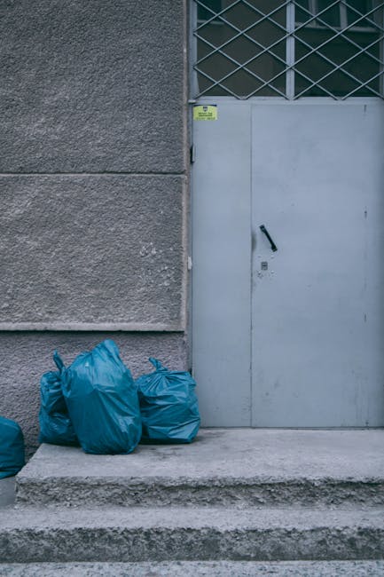 The image shows three blue plastic rubbish bags placed on a concrete doorstep or pavement in front of a grey metal door. The door features a vertical black handle and a small lock, and is set within a building wall made of textured, rough grey concrete. Above the door, there is a metal grille or lattice window with diamond-shaped pattern and dark glass panes. The surrounding environment appears to be an exterior urban setting, possibly part of a commercial or residential property. Natural lighting highlights the textures of the concrete and the plastic bags, with the scene suggesting a private waste collection or disposal area where rubbish is temporarily stored before removal. Waste Clearance Highbury is known for providing independent rubbish removal services, and this scene may reflect an area prepared for scheduled collection or an alternative waste handling approach outside traditional council services.