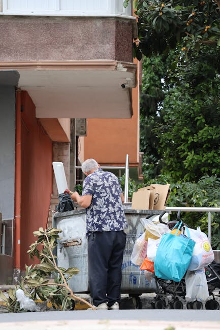 A man with grey hair, wearing a blue and white patterned short-sleeve shirt and dark trousers, is standing outdoors beside a large metal rubbish container on a pavement. He is holding a white plastic bag or container in his right hand, appearing to be either placing or retrieving items from the container. Next to the container, there are several bags of rubbish, including a blue shopping bag, white plastic bags, and a cardboard box, some of which are on a small trolley or cart. The background includes a building with a textured, reddish-brown exterior wall and a small balcony or overhang above him, with lush green foliage of trees visible behind. The scene suggests a typical private waste disposal or rubbish collection activity, consistent with alternative waste handling methods that might be utilized in situations where local authority council rubbish collection access is limited or problematic, in line with the context of on-site clearance and private disposal services provided by Waste Clearance Highbury.