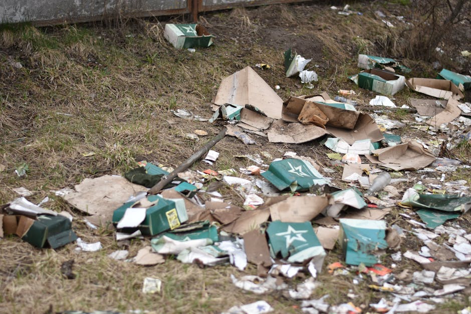 An outdoor scene featuring scattered cardboard boxes and packaging materials on a grassy and dirt area beside a concrete wall, with some boxes torn open and partially flattened. The boxes are primarily green with white branding, and some are crumpled or damaged, revealing contents or inner layers. There are also smaller pieces of paper and plastic debris mixed among the boxes, creating a cluttered appearance. The grass around the litter varies in density, with patches of brown soil visible, and some dried grass and twigs are present. The lighting suggests an overcast day, with diffuse light illuminating the scene evenly. This image exemplifies typical rubbish that might be collected during private or independent waste removal services, such as those offered by Waste Clearance Highbury, especially when dealing with unsheltered or scattered rubbish beyond regular council collection coverage.