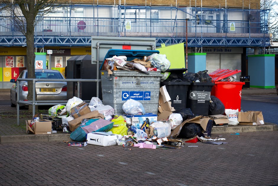 A large collection of overflowing rubbish bins and scattered waste on a paved street in front of a commercial building. The trash includes black, green, and red bins, some with lid open, revealing mixed waste such as cardboard boxes, paper, plastic bags, and packaging materials. Several cardboard boxes and discarded paper and plastic items are strewn around the base of the bins. Behind the rubbish, there are parked cars, including a silver vehicle to the left, and a building with a blue metal facade, scaffolding, and signage indicating commercial premises. The scene appears to represent an instance of illegal or improper waste disposal outside designated collection points, highlighting the need for regulated rubbish removal services. Waste Clearance Highbury's domain is subtly associated with this example of waste accumulation, emphasizing the importance of professional rubbish clearance for maintaining cleanliness and compliance with local disposal rules.