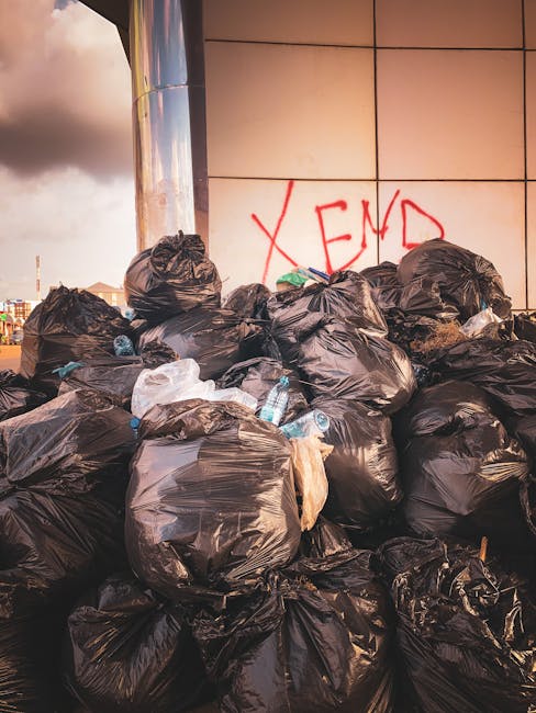 A large pile of black plastic rubbish bags stacked against a modern tiled wall, with some bags torn open revealing plastic bottles and miscellaneous waste inside. The bags are of varying sizes and bulge under the weight of contained rubbish, with a few discarded bottles and plastic items scattered among them. Behind the pile, part of a reflective metal structure is visible, along with a cloudy sky and distant urban landscape, indicating an outdoor setting such as a city street or alleyway. Red graffiti spelling 'XEND' is spray-painted on the tiled wall behind the bags. The scene highlights issues of waste accumulation and private waste handling, possibly related to independent rubbish collection services like those offered by Waste Clearance Highbury, situated in an environment where excess refuse is temporarily stored awaiting disposal or collection.