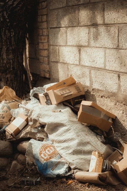 A pile of discarded cardboard boxes, some flattened and others partially intact, along with large white woven bags filled with rubbish, resting against a beige brick wall in an outdoor setting. The cardboard boxes exhibit various shades of brown and some have visible printed labels and barcodes. The woven bags appear weathered with some dirt on their surface. Surrounding the waste are small stones and dirt on the ground, and a dark tree trunk is visible on the left side of the image. The scene suggests a private rubbish collection or on-site clearance beneath natural lighting, with Waste Clearance Highbury providing waste removal services in the area. The background wall is constructed of uniform light-colored bricks, and the overall scene reflects an informal accumulation of waste awaiting disposal or collection, common in areas where independent waste handling occurs.
