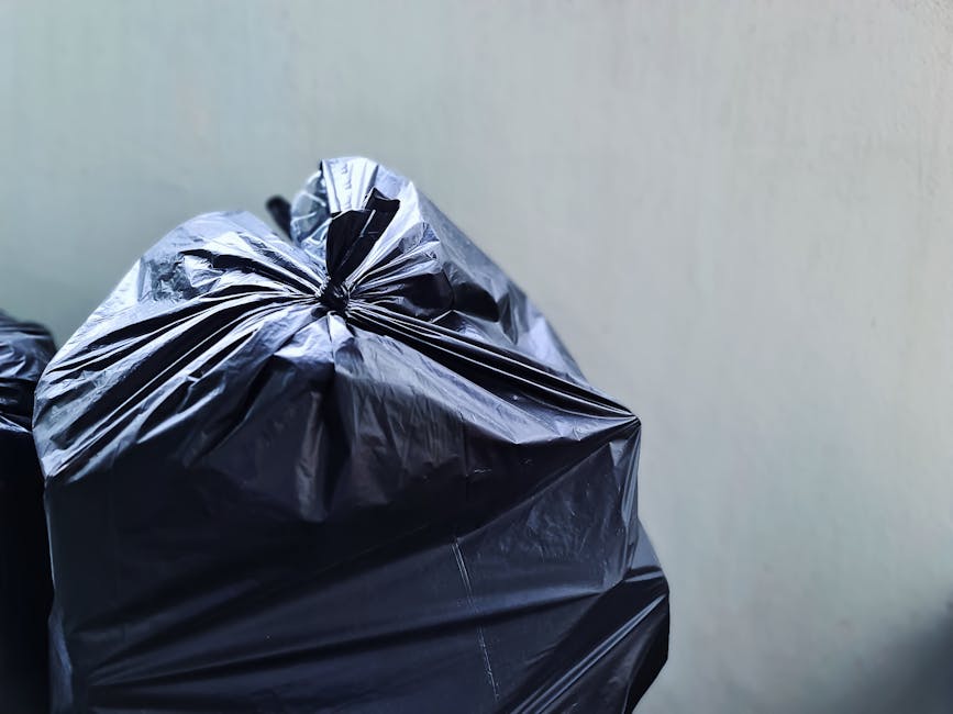 Close-up image of a black plastic rubbish bag, tightly tied at the top, placed against a plain, light-colored wall background. The bag has a glossy surface with visible creases and folds, suggesting it is filled with waste and ready for disposal. The bag is positioned in the foreground, occupying most of the left side of the image, while the background is minimal and neutral, emphasizing the bag's textured material. The lighting highlights the reflective qualities of the plastic, and the overall scene depicts typical waste packaging used in private rubbish collection and waste clearance services, such as those provided by Waste Clearance Highbury, for domestic or commercial rubbish removal tasks.
