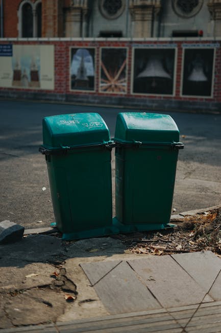 Two green plastic rubbish bins with black lids are positioned side by side on a concrete sidewalk near the edge of a street. The bins are slightly weathered, with visible scratches and marks on their surface, and are placed on a small black plastic mat that helps to stabilize them. The setting is an urban area with a paved road behind the bins, which appears to be busy and well-used, with a slight sheen from recent rain or vehicle traffic. In the background, a brick wall with advertising posters and display windows for a shop or restaurant is visible, featuring images of curtains or drapes. The sidewalk is made of concrete slabs, with some loose and uneven tiles near the base of the bins, and fallen leaves and small debris scattered around. The overall scene reflects typical on-street waste collection suited for private or independent rubbish disposal services, highlighting the importance of well-placed waste management solutions in public urban spaces, as offered by waste clearance specialists like Waste Clearance Highbury.