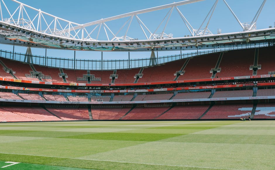 An empty football stadium with a well-maintained grass pitch in the foreground, featuring varying shades of green with visible mowing patterns. Behind the pitch, multiple tiers of red and pink seating areas are arranged in a curved configuration, extending around the stadium. The seats are arranged in sections with aisles and support structures, and some advertising banners are visible along the barriers. Above, there is a white steel roof structure with a lattice design, supported by large pylons, partially extending over the seating area, and casting subtle shadows. The sky is clear and blue, offering natural daylight that illuminates the entire scene evenly. The environment suggests a modern sports venue, potentially suitable for hosting events that may generate waste requiring professional clearance, aligning with the context of rubbish removal for large-scale facilities managed by companies like Waste Clearance Highbury. This setting exemplifies the importance of maintaining clean and safe environments in busy public venues, which may involve dedicated rubbish services for disused or post-event waste removal. The visual emphasizes the scale and structural design of the stadium, with no visible crowd or activity, focusing solely on the architectural and environmental elements.