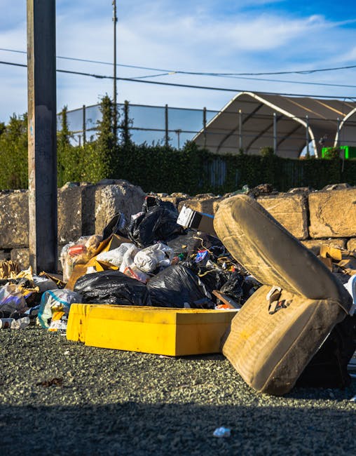 A close-up view of a large pile of discarded rubbish and waste materials on a gravel surface, including black plastic bags, a yellow plastic container, and an old, worn upholstered chair seat or cushion with a fabric cover. The rubbish is cluttered and appears to contain general household waste. In the background, there is a low stone wall made of irregularly shaped, rough-textured stones, with some greenery, including bushes and trees, behind it. Overhead, power lines stretch across the partly cloudy sky, which is blue with a few wispy clouds. Behind the wall, a large, semi-circular metal structure with a protective cover, possibly part of a sports or event facility, is visible. The scene suggests an area designated for rubbish collection, possibly linked to private waste removal services, and highlights the significance of proper waste disposal, aligning with the themes of independent rubbish clearance.