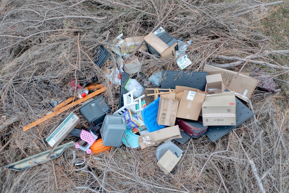 A large pile of assorted household and electronic waste materials situated outdoors on a patch of dry, brown grass and thin, leafless branches. The collection includes cardboard boxes, some flattened and others still partially assembled, with visible printed labels and tape, alongside plastic storage containers in shades of gray and blue; several black plastic trays and trays with ridges are also present. There are various small objects, such as a pink toy or tool, a red soda can, and a green and black handheld device, all mixed within the debris. Additionally, a wooden and black metal piece possibly part of furniture or equipment leans among the clutter. The waste appears haphazardly dumped, with some items stacked atop each other, creating a disorganized heap that contrasts sharply with the natural environment surrounding it. This scene relates to private waste disposal or rubbish removal services, such as those offered by Waste Clearance Highbury, which handles on-site clearance of unwanted household debris outside of regular council collections. The lighting is natural, indicating daylight, and no activity or people are visible in the scene, emphasizing the outdoor context of the waste accumulation.