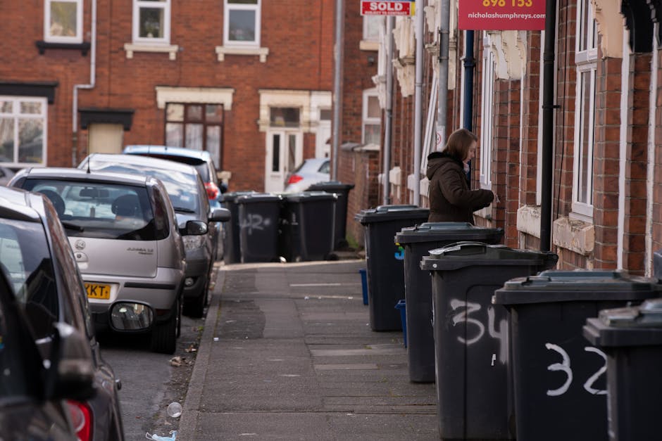 A woman stands on a paved sidewalk beside several large black wheelie bins with closed lids, positioned against the brick exterior wall of a residential building on Holloway Road in Highbury. The bins are arranged in a neat row, each with white painted numbers on their sides, indicating designated waste containers. The woman, dressed in a dark jacket and trousers, appears to be handling or retrieving items from a bin or nearby. The background features parked cars along the curb, including a silver hatchback closest to the camera, with additional vehicles further down the street. The building's brick facade has white window frames and is partially obscured by a sign advertising a property sale, and another sign for a waste collection service is visible on the building. The scene occurs during daylight with natural, slightly overcast lighting, capturing an everyday urban environment where private waste disposal methods are evident through the presence of multiple rubbish bins along the pavement, illustrating a typical street scene related to rubbish collection and waste management services in the area.