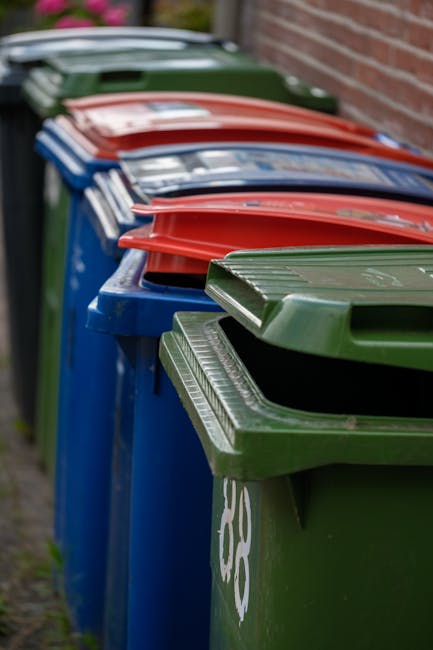 A row of large, heavy-duty wheelie bins lined up along a pavement beside a brick wall, with the bins in various colours including green, blue, red, and black. The green bin in the foreground has a textured lid with parallel grooves, and the blue bins behind it feature similar design elements. The red-lidded bin is positioned behind the blue ones, with its lid slightly ajar. The plastic bins are used for waste collection and are arranged in a neat line, suggesting they are awaiting emptying or for private waste disposal services. The scene is set outdoors during daylight, with natural light illuminating the bins, which are situated directly on the pavement adjacent to a building's brick wall, emphasizing the everyday urban environment typical of waste management in residential areas.