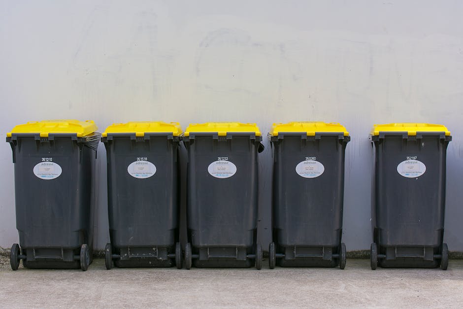 A woman stands on a paved sidewalk beside several large black wheelie bins with closed lids, positioned against the brick exterior wall of a residential building on Holloway Road in Highbury. The bins are arranged in a neat row, each with white painted numbers on their sides, indicating designated waste containers. The woman, dressed in a dark jacket and trousers, appears to be handling or retrieving items from a bin or nearby. The background features parked cars along the curb, including a silver hatchback closest to the camera, with additional vehicles further down the street. The building's brick facade has white window frames and is partially obscured by a sign advertising a property sale, and another sign for a waste collection service is visible on the building. The scene occurs during daylight with natural, slightly overcast lighting, capturing an everyday urban environment where private waste disposal methods are evident through the presence of multiple rubbish bins along the pavement, illustrating a typical street scene related to rubbish collection and waste management services in the area.